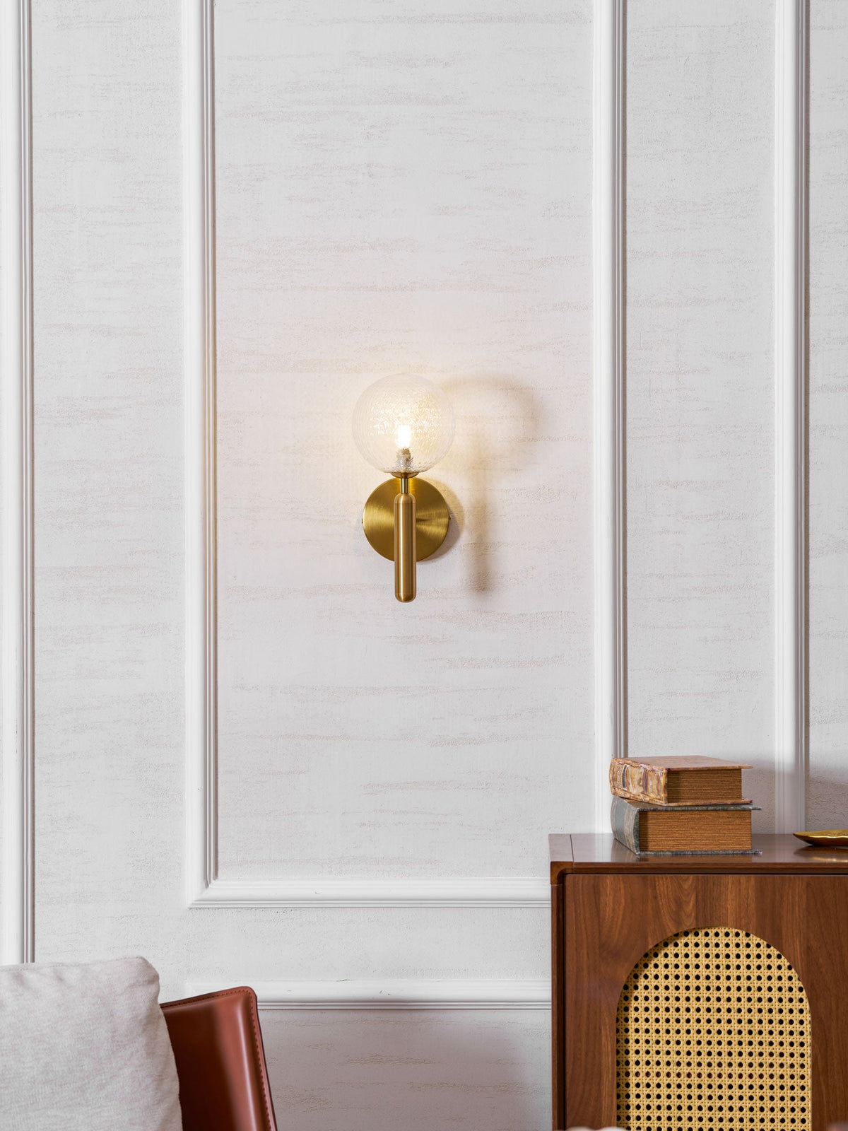 Beside a wood cabinet with books, this brass Clérachic wall light's clear glass globe glows on a panelled wall.