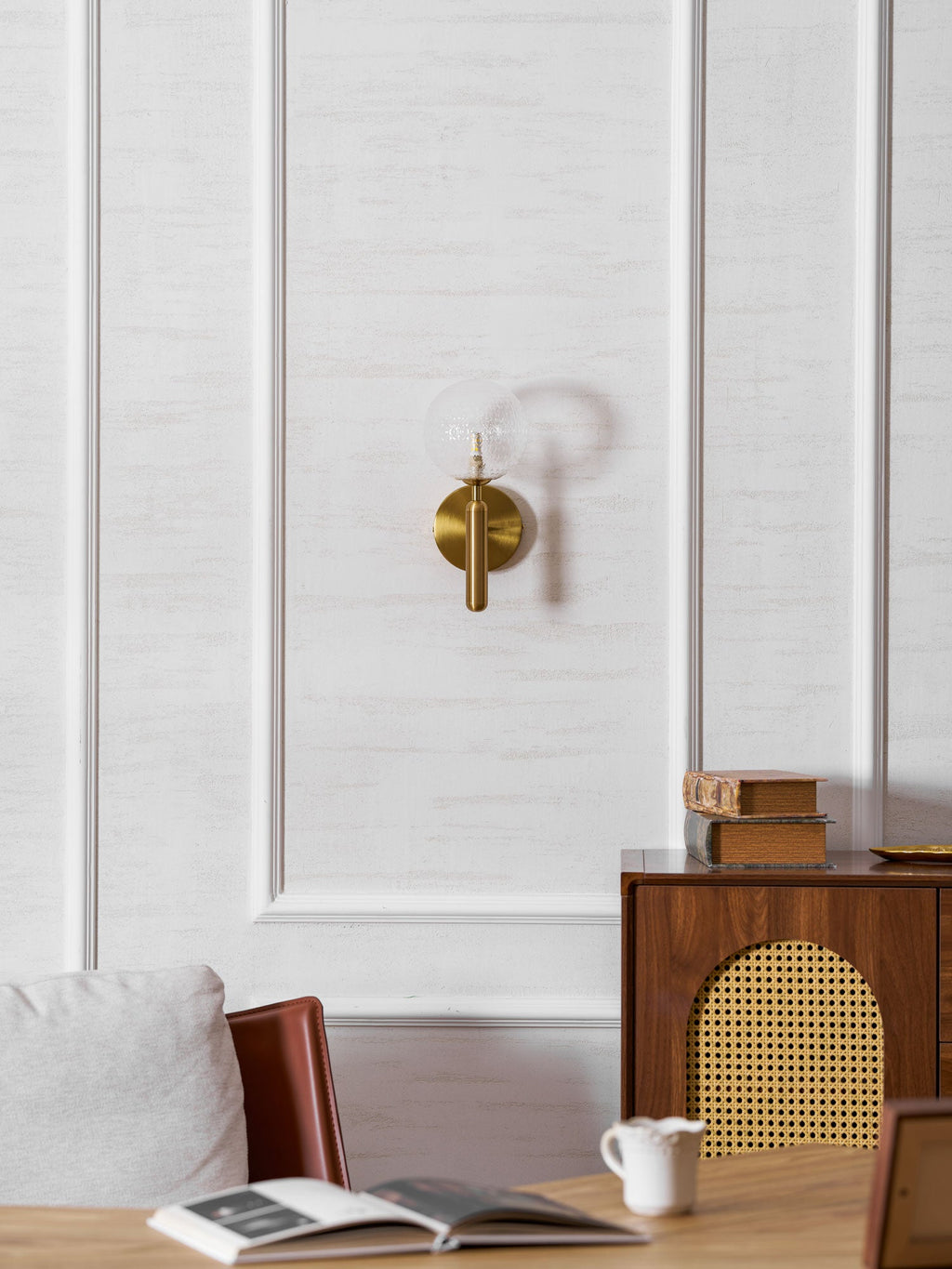 In a wider view with a desk, chair and open book, this brass Clérachic wall light's clear glass globe glows on a panelled wall.