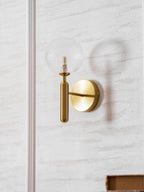 Near a book's corner, this brass Clérachic wall light's clear glass globe glows on a panelled wall.