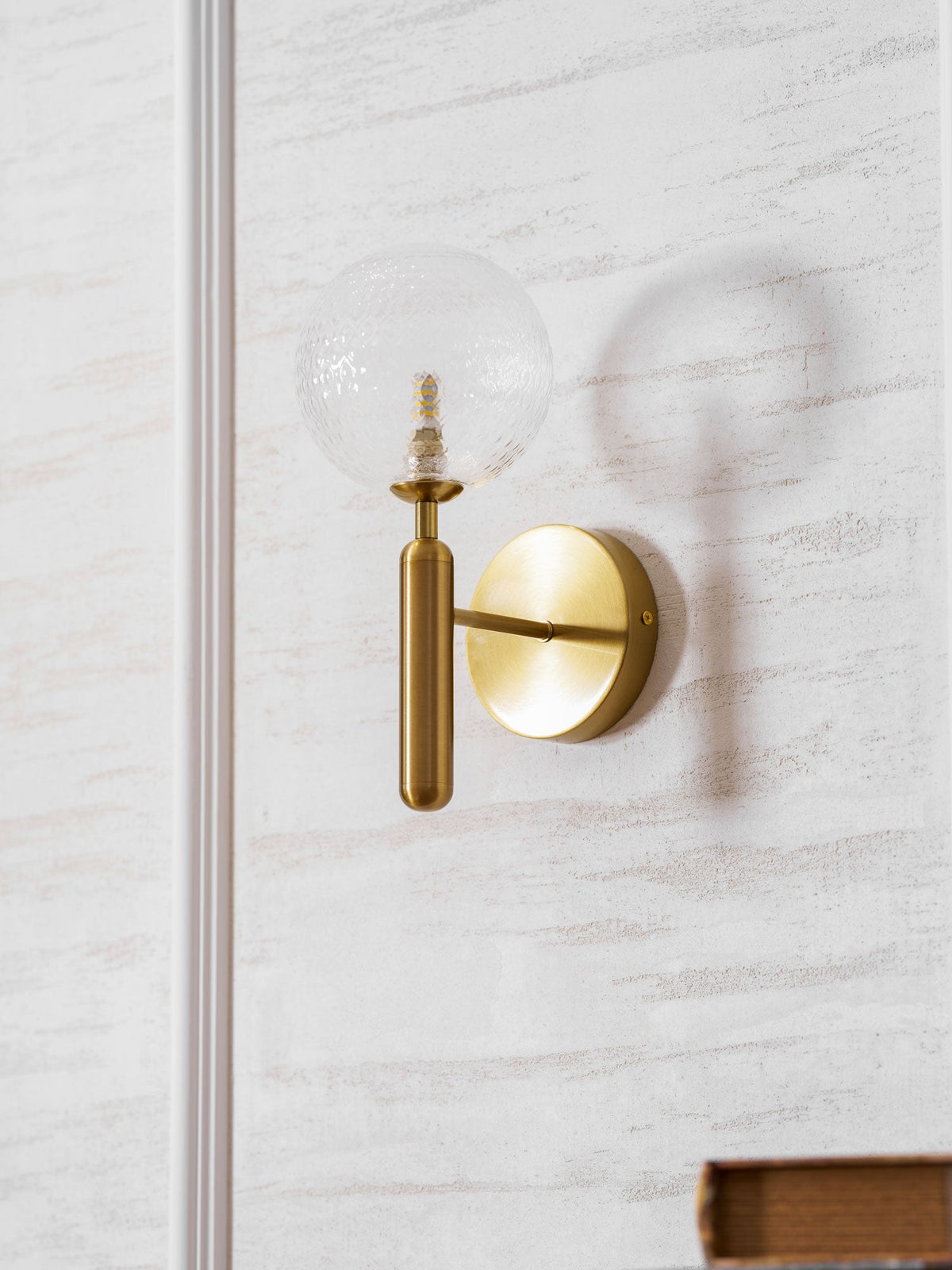 Near a book's corner, this brass Clérachic wall light's clear glass globe glows on a panelled wall.