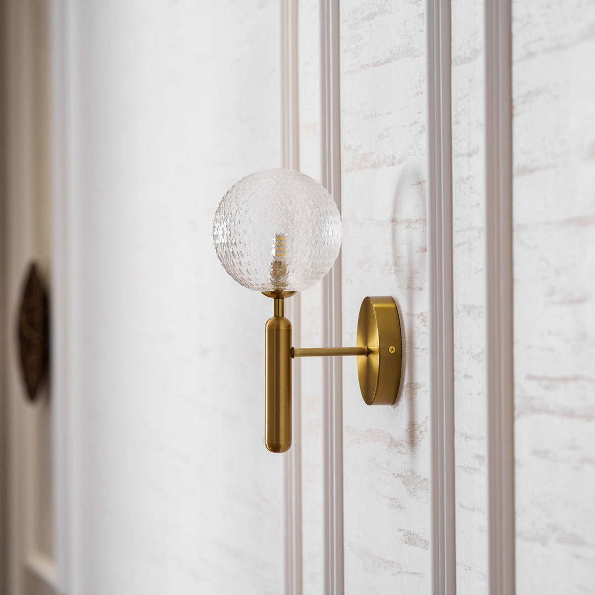 Close to a door edge, this brass Clérachic wall light's dimpled clear glass globe glows on a panelled wall.