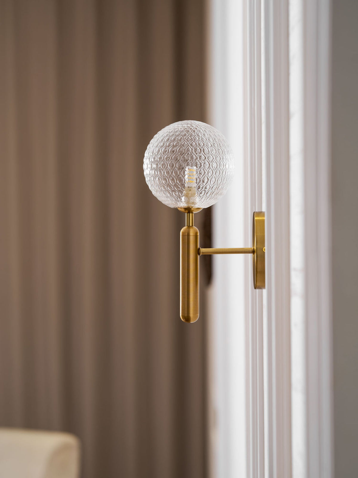 In a warm-toned hallway, this brass Clérachic wall light's dimpled clear glass globe glows beside a wooden door.