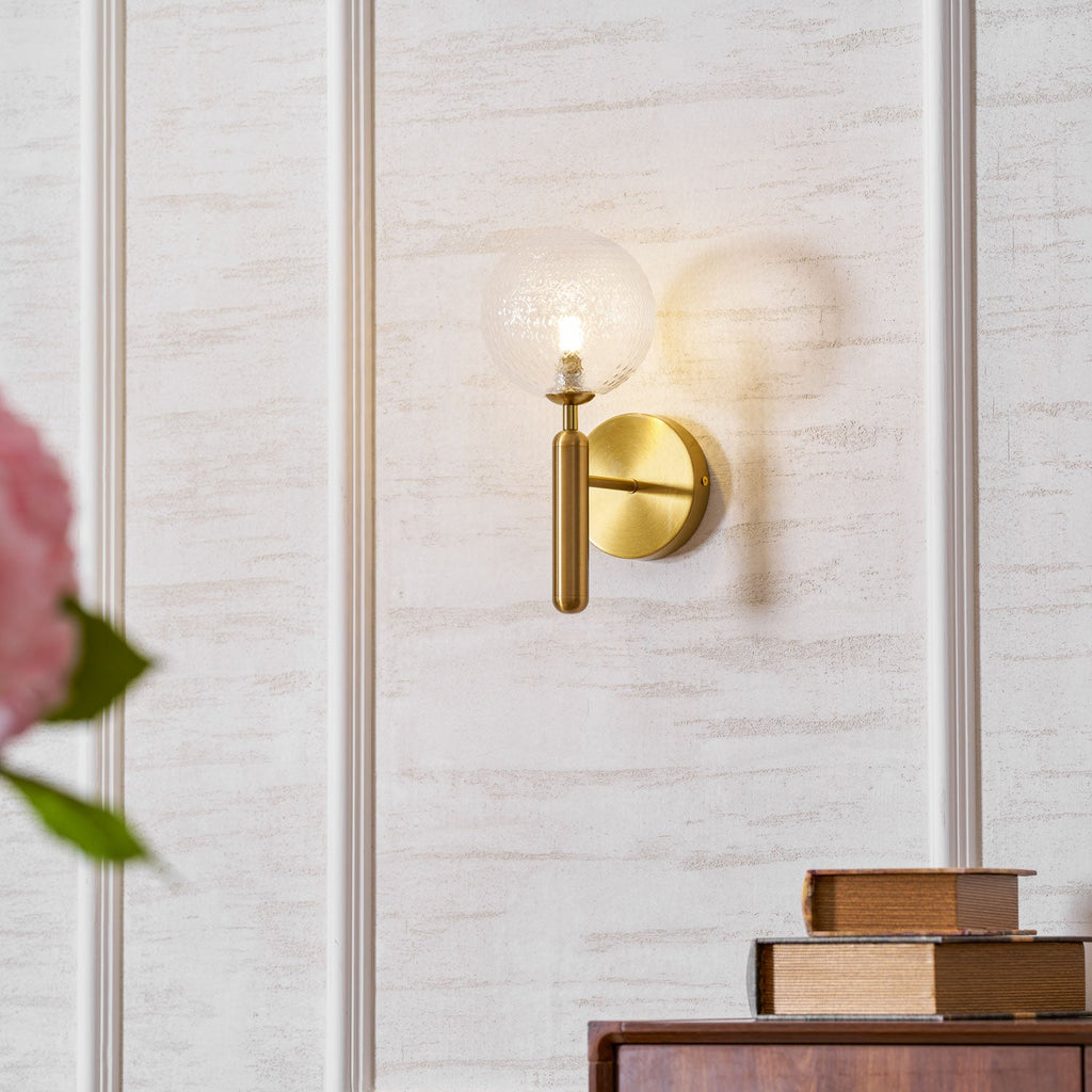 Beside a pink flower and a stack of books, this brass Clérachic wall light's clear glass globe glows against a plaster-textured wall.