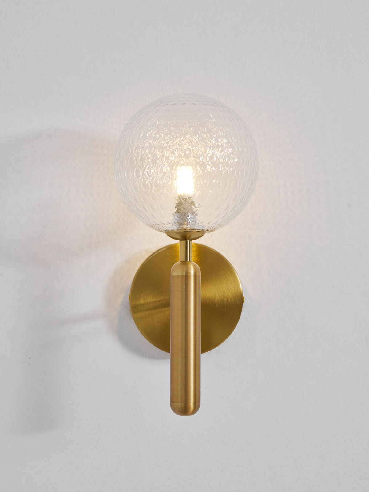 This brass Clérachic wall light's dimpled clear glass globe glows softly, its bulb visible, on a light grey textured wall.