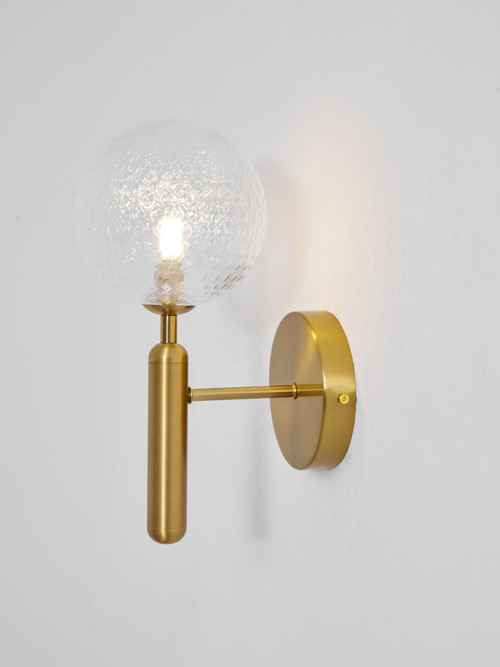 Seen from the side, this brass Clérachic wall light's lit dimpled clear glass globe glows on a light grey wall.