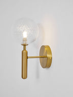 Seen from the side, this brass Clérachic wall light's lit dimpled clear glass globe glows on a light grey wall.