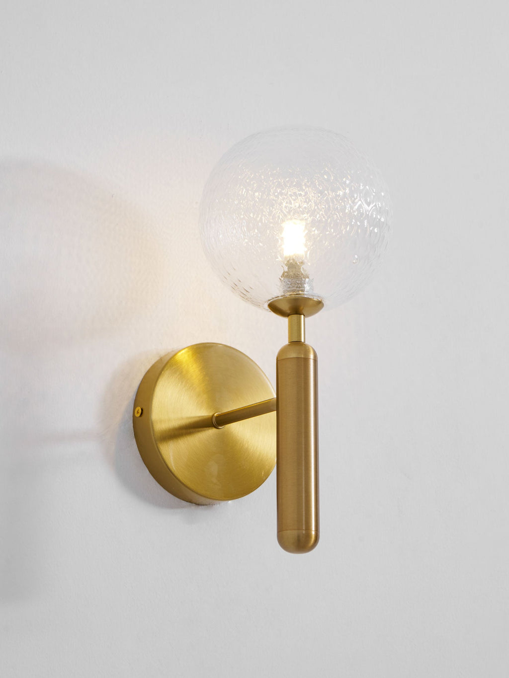 Viewed from the left, this brass Clérachic wall light's dimpled clear glass globe glows on a light grey wall.
