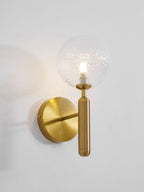 Viewed from the left, this brass Clérachic wall light's dimpled clear glass globe glows on a light grey wall.