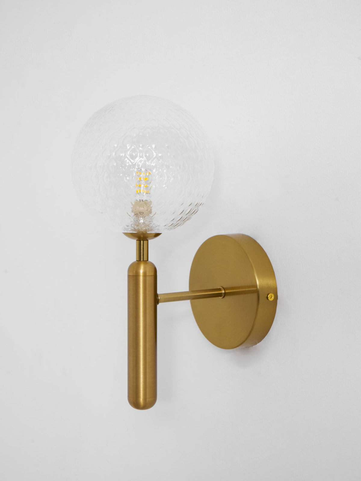 This brass Clérachic wall light's dimpled clear glass globe glows in close-up against a light grey wall.