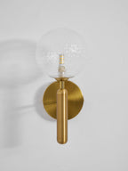 Facing forward, this brass Clérachic wall light's dimpled clear glass globe glows on a light grey wall.