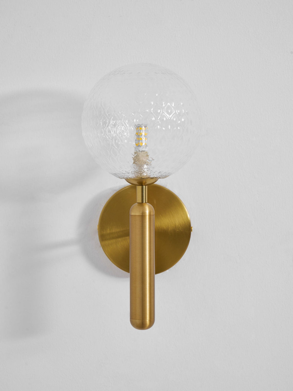 Facing forward, this brass Clérachic wall light's dimpled clear glass globe glows on a light grey wall.