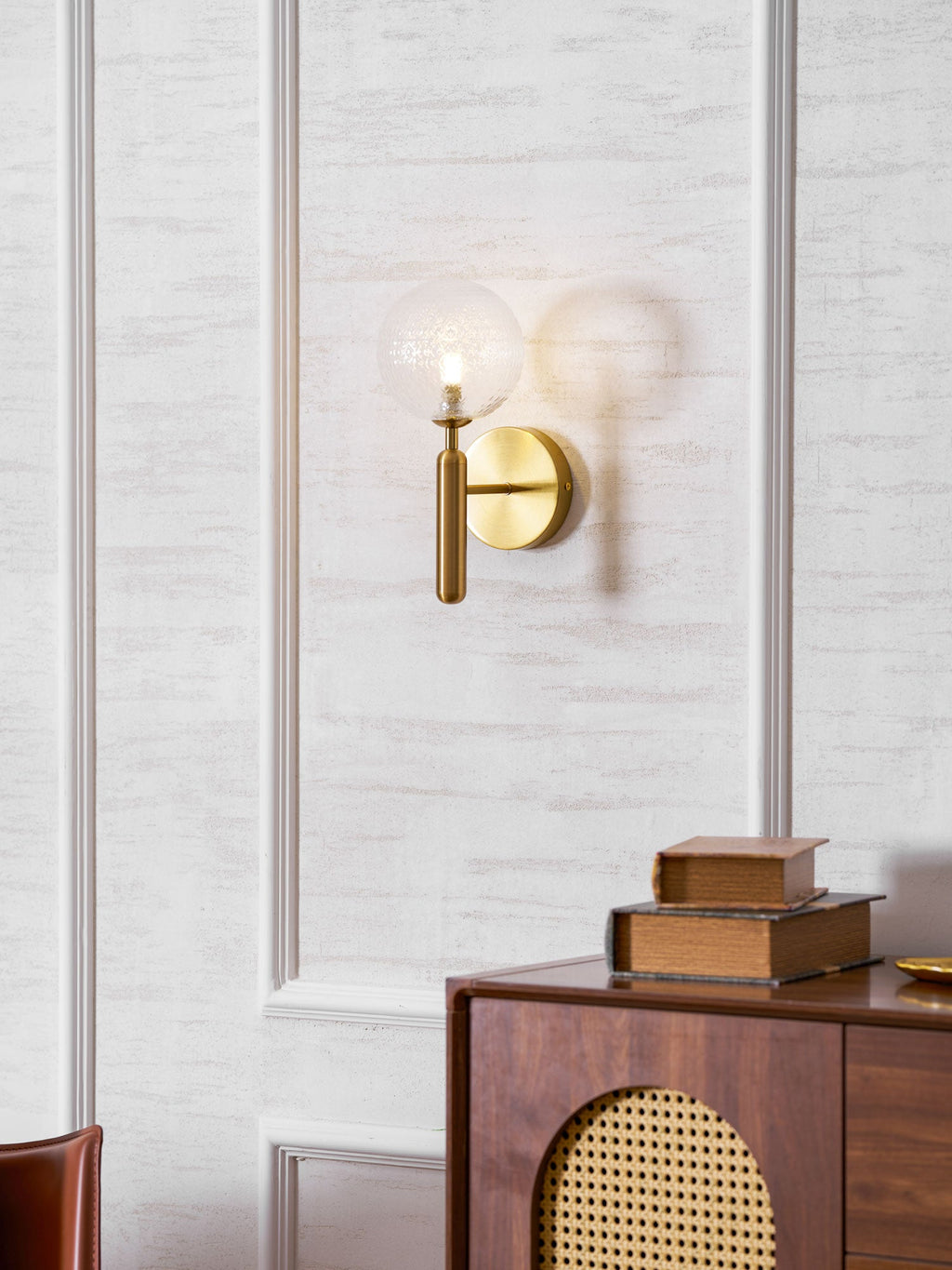 Softly out of focus against a panelled wall with books and a rattan-fronted cabinet nearby, this brass Clérachic wall light's glass globe glows warmly.