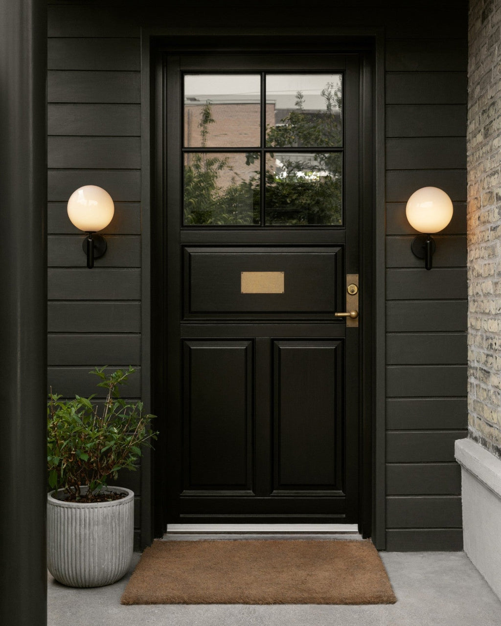 Flanking a black panelled front door with a brass mail slot, two black Clérachic wall lights glow above a doormat.