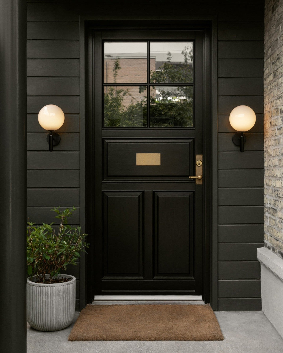 Flanking a black panelled front door with a brass mail slot, two black Clérachic wall lights glow above a doormat.