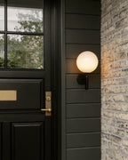 Beside a black door framed in brick, this black Clérachic wall light glows with its round white glass globe.