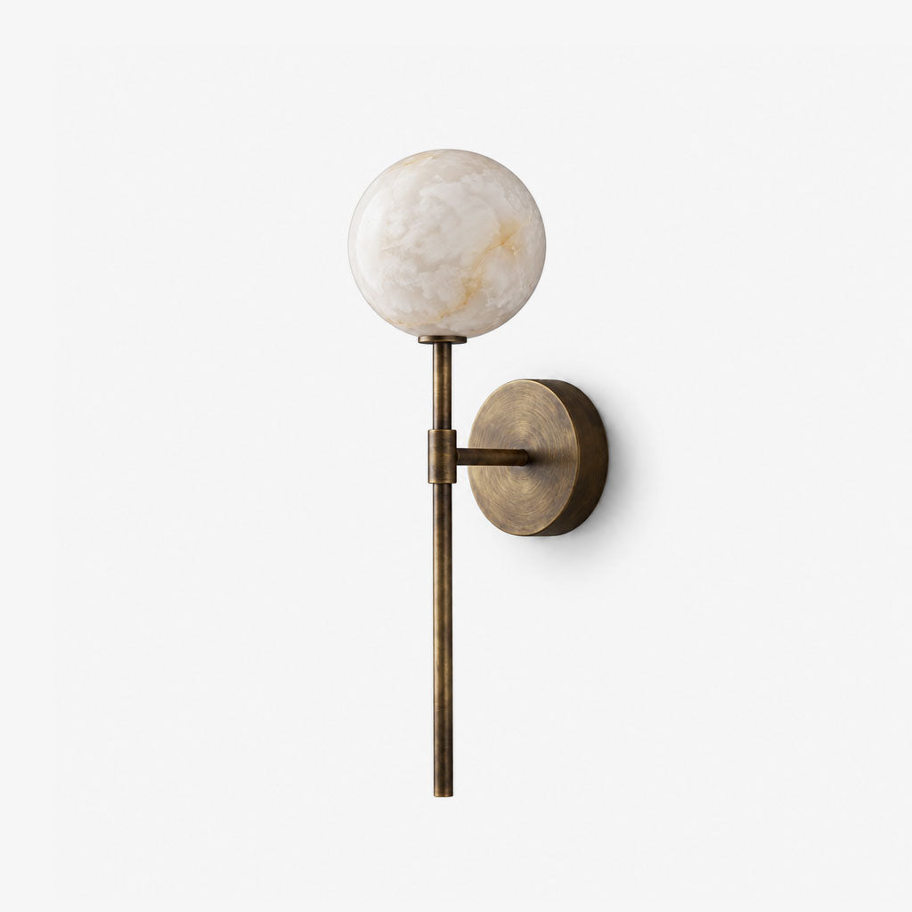 This Clérachic wall sconce in brass stands alone against a plain white backdrop, its ball shade lit.