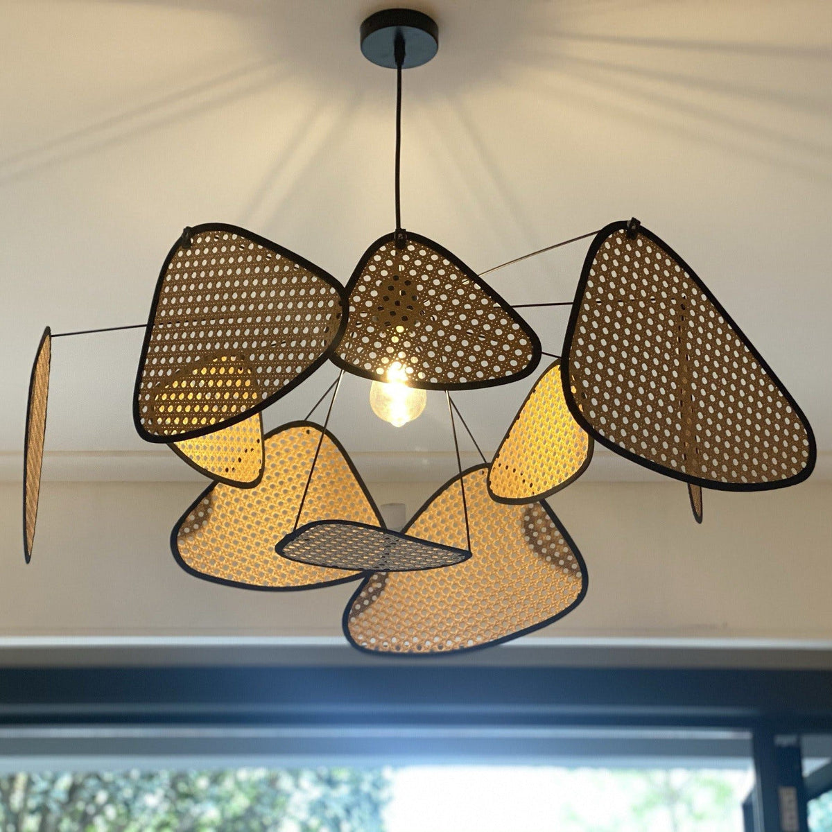 Greenery is visible through a tall window behind this Clérachic pendant, its bulb glowing beneath dark and tan paddles.