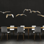 White bird-shaped pendants from this Clérachic fixture hang above a long table with grey chairs, on a dark backdrop.