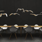 A cluster of white bird pendants from this Clérachic light hangs above a long wooden table with white chairs, on a dark backdrop.