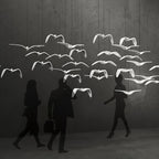 A flock of white bird pendants from this Clérachic light hangs above walking figures silhouetted against a grey backdrop.