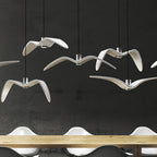 White bird-shaped pendants from this Clérachic fixture hang low above a wooden table, seen close against a dark backdrop.
