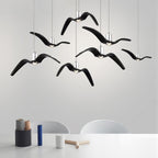 Black bird-shaped pendants from this Clérachic fixture hang above a table set with vases and decor, against a light-grey wall.