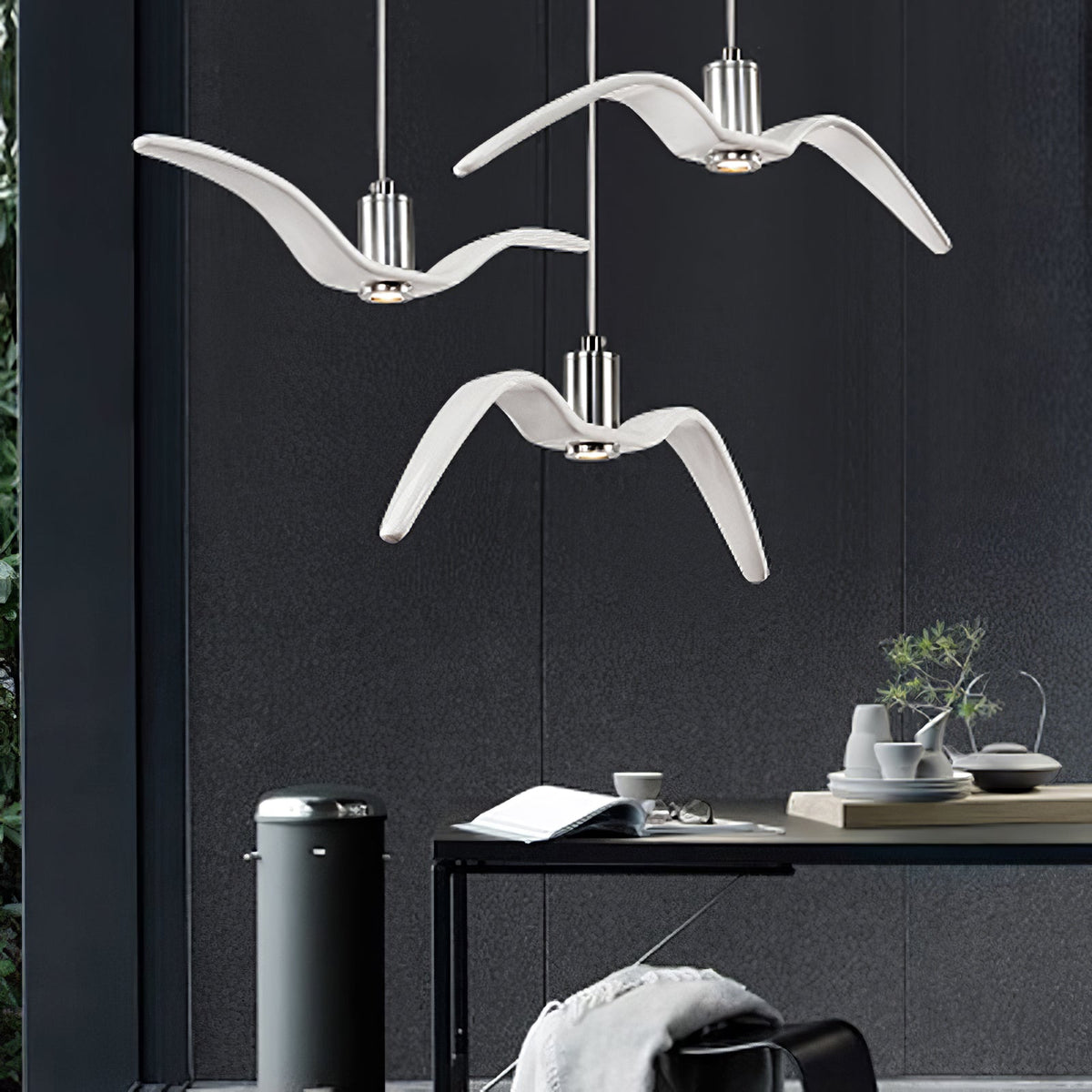 Three white bird-shaped pendants from this Clérachic fixture hang at staggered lengths above a home office desk with a bin and plant.