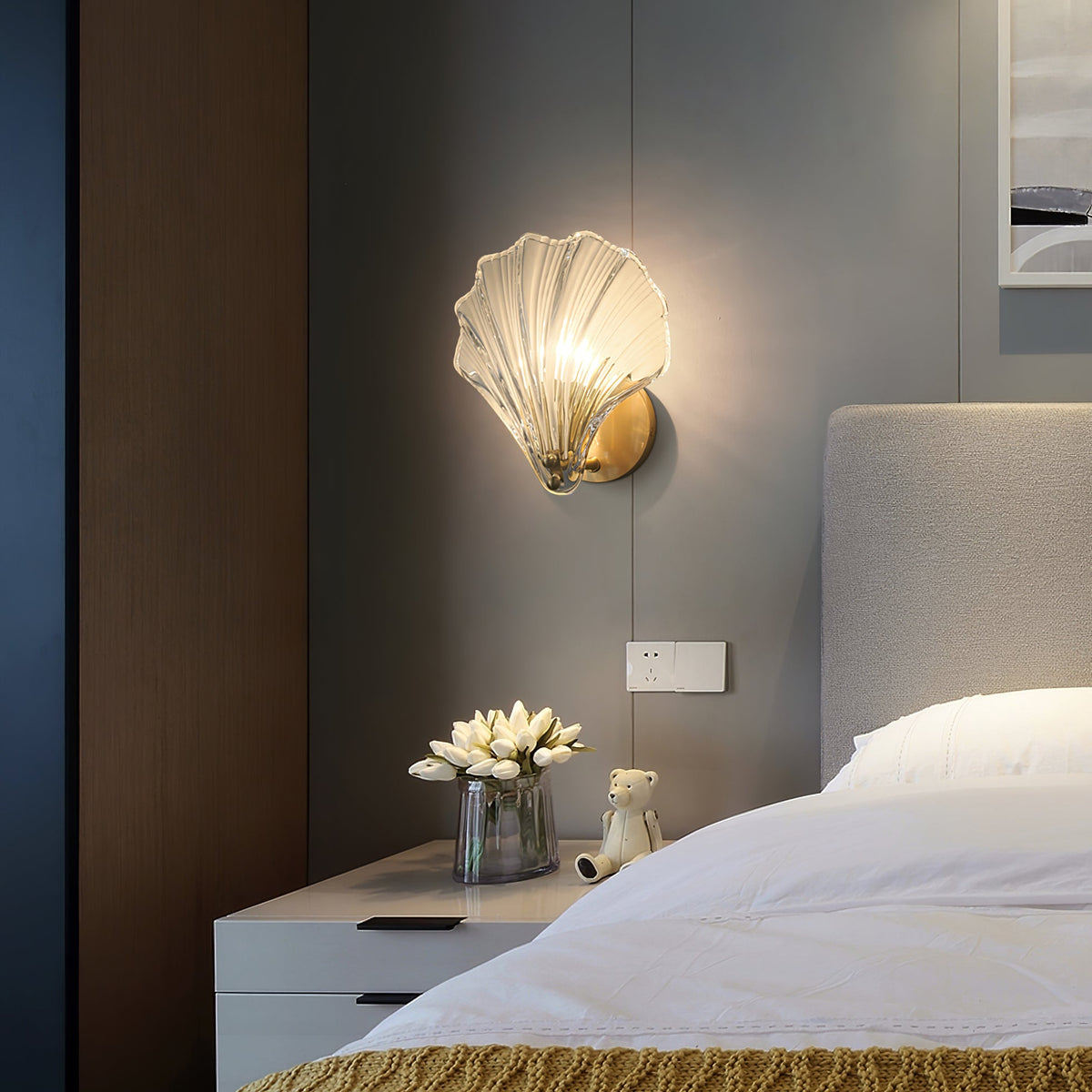 In a dark, moody bedroom, this Clérachic wall light glows amber above a nightstand set with white flowers.