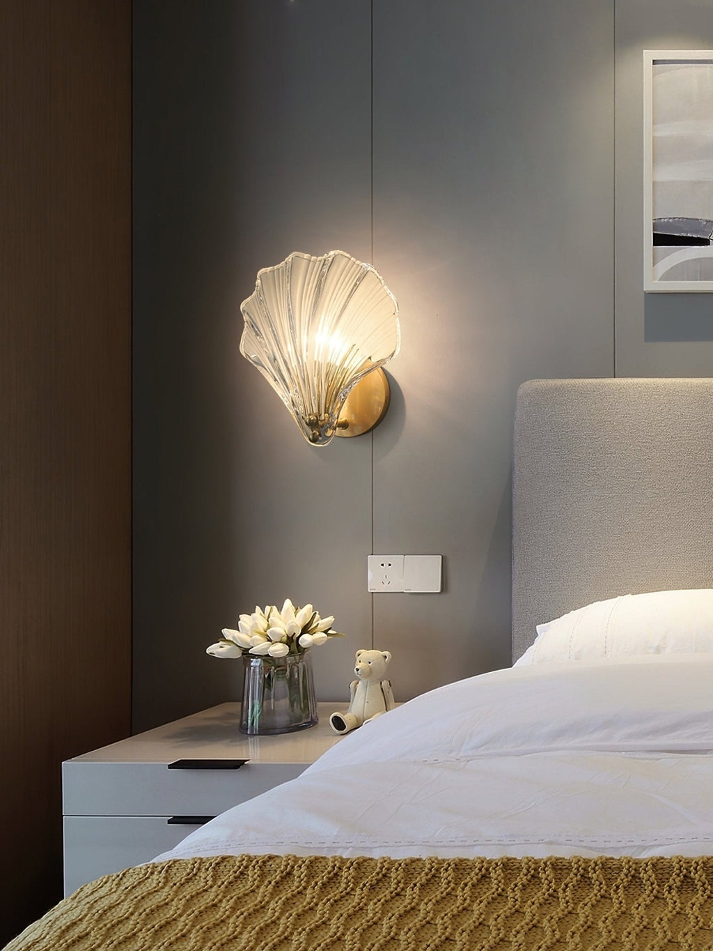 Mounted above a nightstand with tulip flowers, this Clérachic wall light glows beside an upholstered headboard.