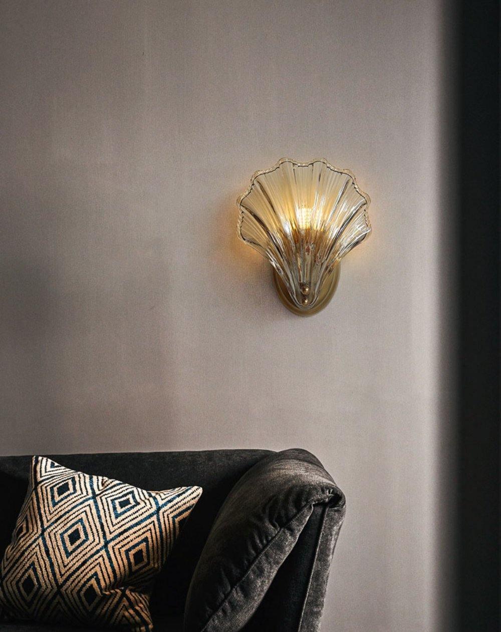 Beside a dark cushioned sofa with a patterned throw pillow, this Clérachic wall light glows against a shadowed wall.