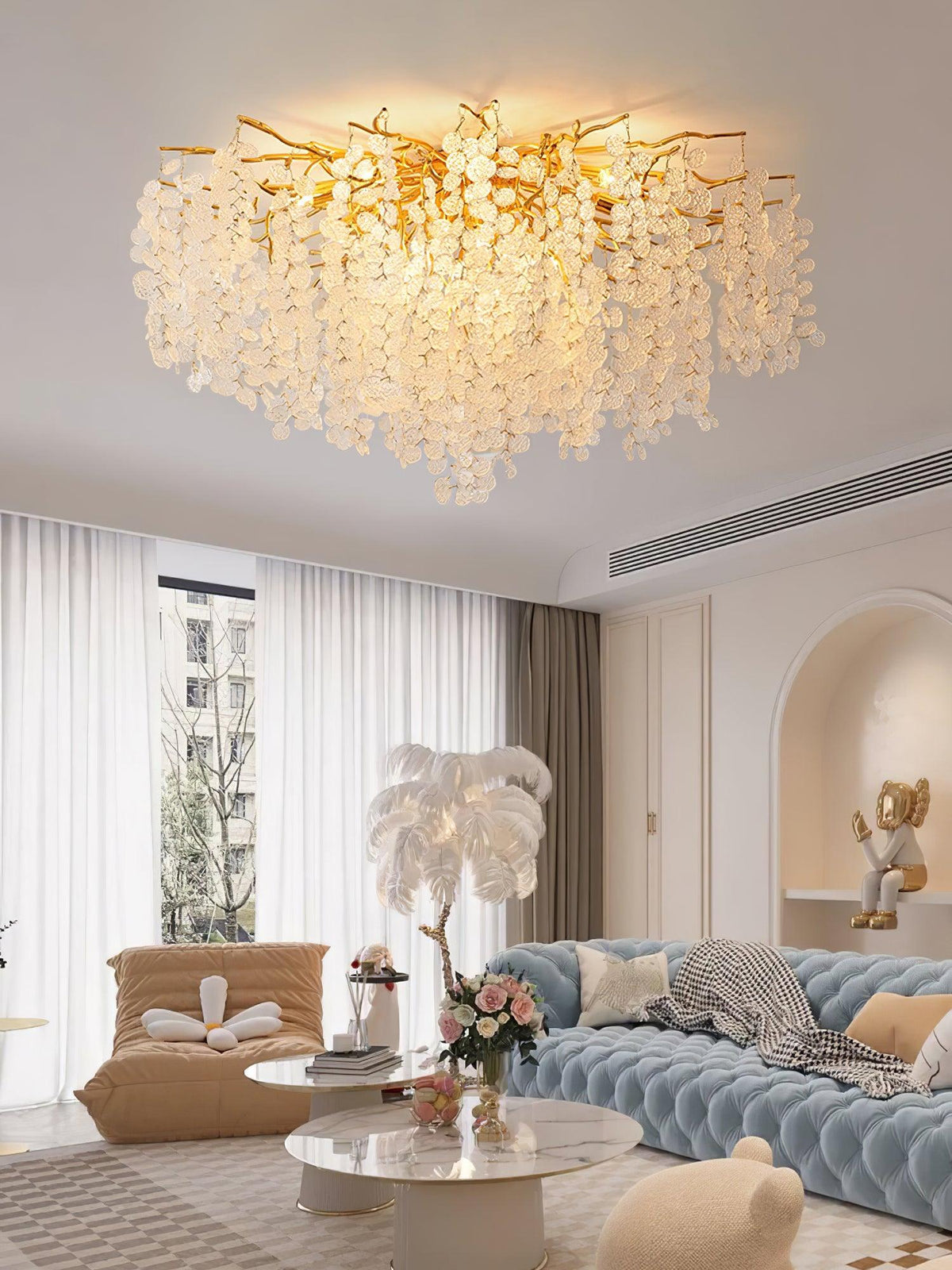 Beside a feather branch arrangement, this Clérachic ceiling light glows above a blue tufted sofa in a living room with sheer curtains.