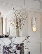 Reflected in a mirror above a veined marble fireplace, the Clérachic wall sconce lights a vase of bare branches.