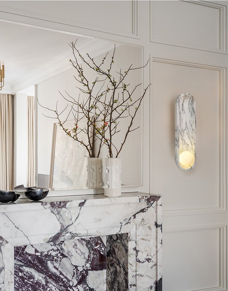 Reflected in a mirror above a veined marble fireplace, the Clérachic wall sconce lights a vase of bare branches.