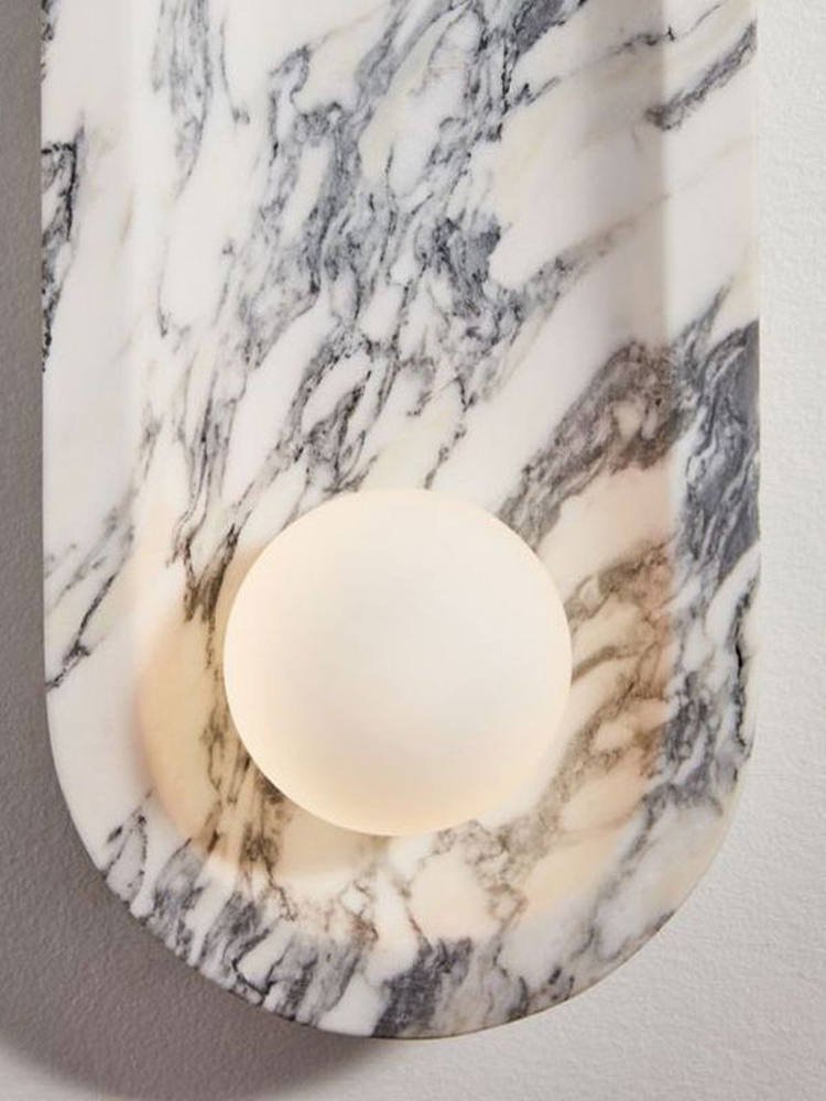 A close-up on the Clérachic sconce's frosted opal sphere glowing warmly within its grey-veined marble niche.