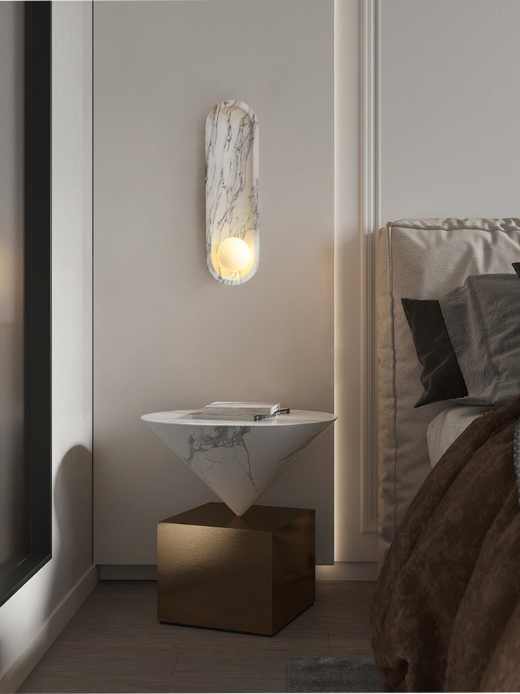 By a window in a bedroom corner, the lit Clérachic wall light sits above a cone-shaped table on a brass base.