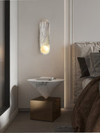 By a window in a bedroom corner, the lit Clérachic wall light sits above a cone-shaped table on a brass base.