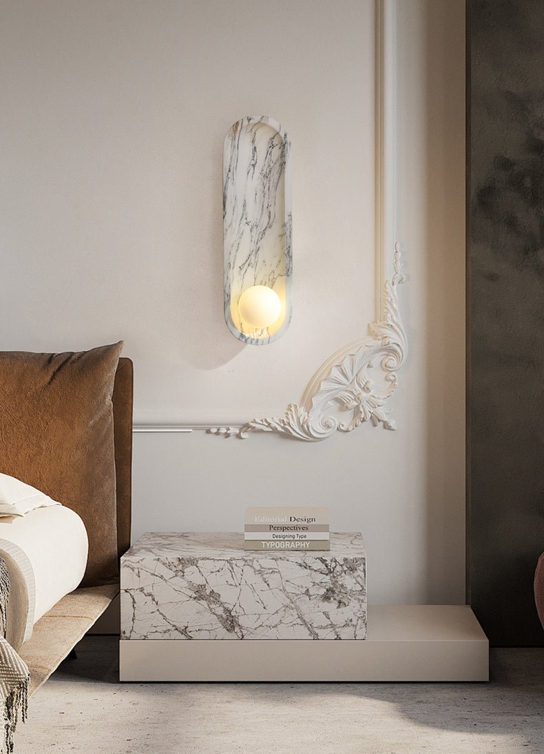 Mounted above a marble bedside plinth, this Clérachic wall light warms a bedroom corner beside an ornate mirror edge.