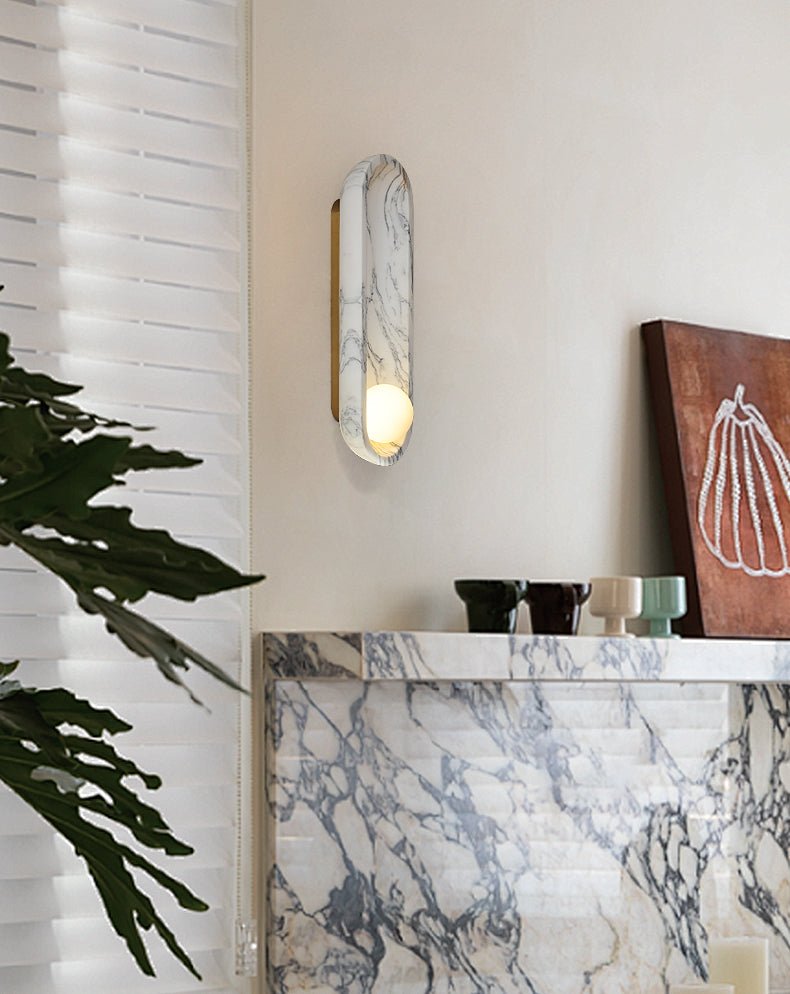 The Clérachic wall sconce lights a marble mantelshelf holding glassware and a trailing green plant beside framed art.