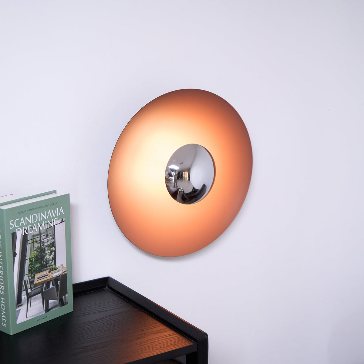 Lit orange disc wall light with a chrome centre from Clérachic, mounted above a dark wood cabinet holding a magazine.