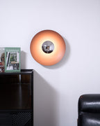 This orange Clérachic wall light glows softly above a dark cabinet, its chrome centre catching the light, beside an armchair.