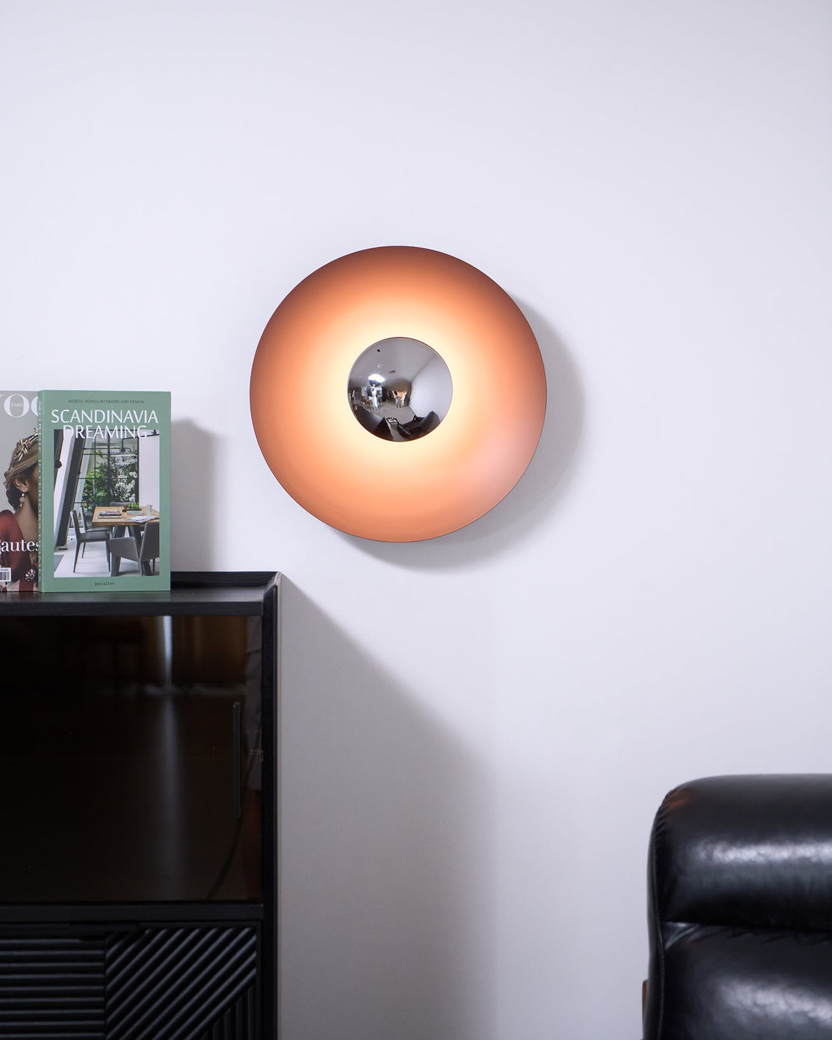 This orange Clérachic wall light glows softly above a dark cabinet, its chrome centre catching the light, beside an armchair.