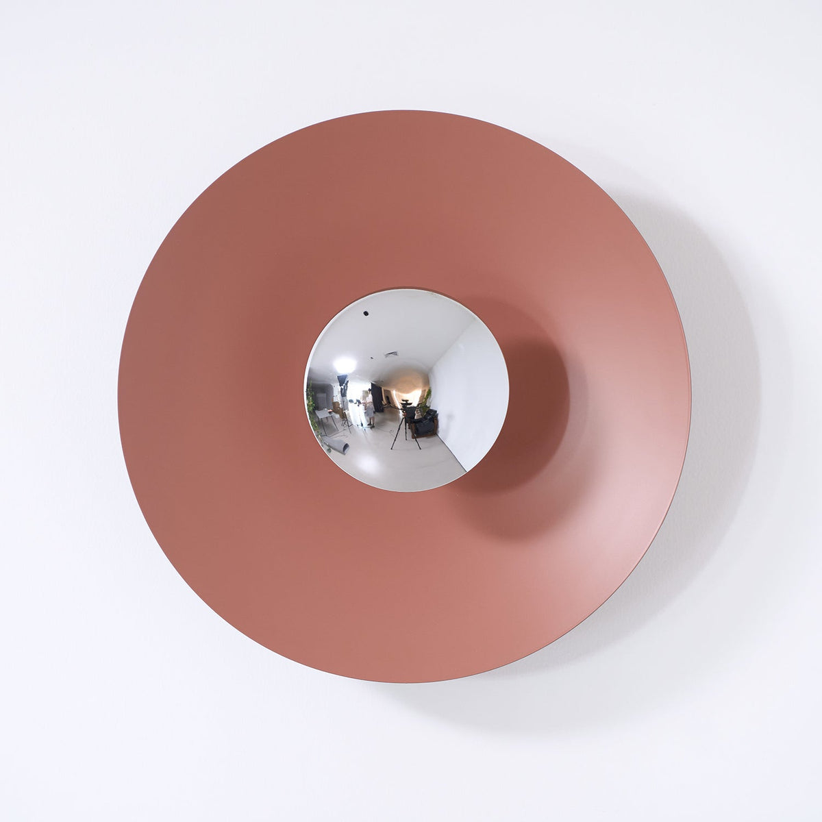 A dusty-rose Clérachic wall light on a plain wall, its mirrored centre reflecting the camera equipment.