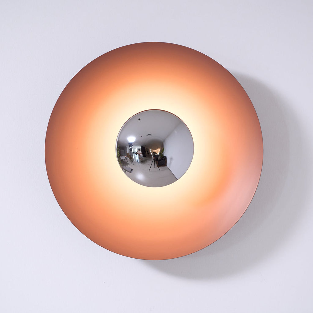 This dusty-rose Clérachic wall light glows warm on a plain wall, its mirrored centre reflecting the studio.