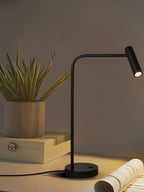 An olive-green wall sits behind this black Clérachic table lamp, lit, beside a potted plant.