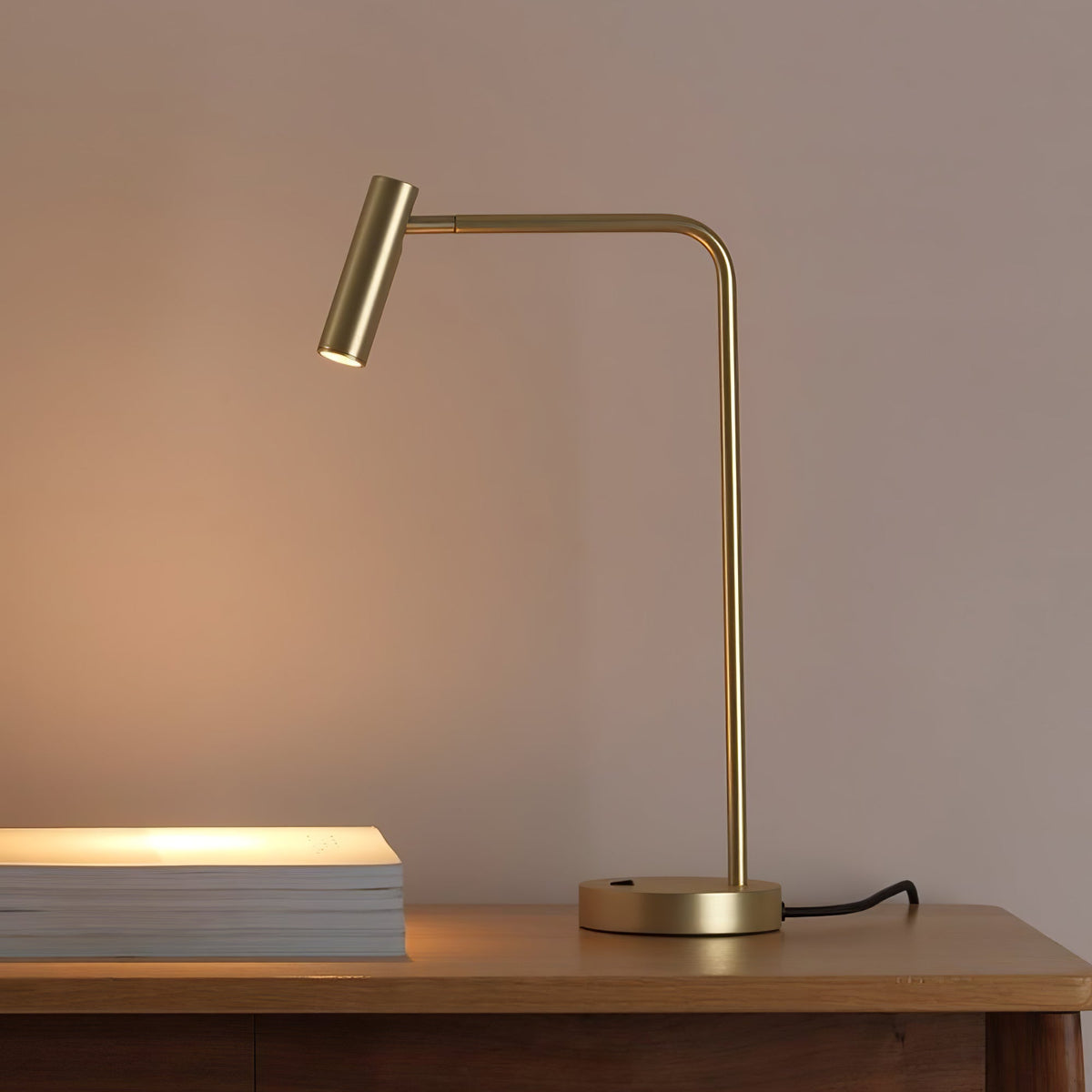 A stack of white books sits below this gold Clérachic table lamp, lit, on a wood table.