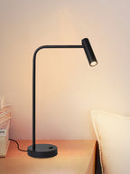 A white pillow sits behind this black Clérachic table lamp, lit, on a wood nightstand.
