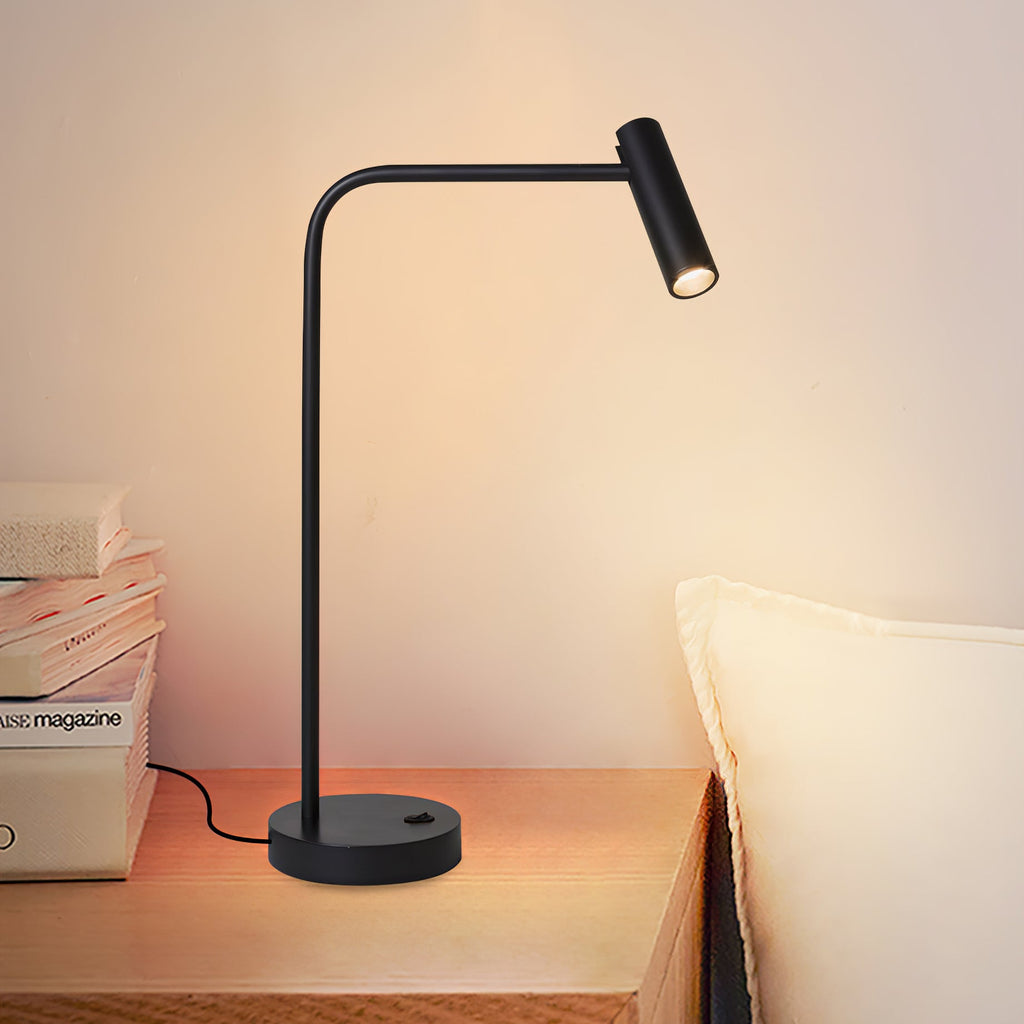 Stacked magazines and a pillow sit beside this Clérachic table lamp, lit, on a wood nightstand.