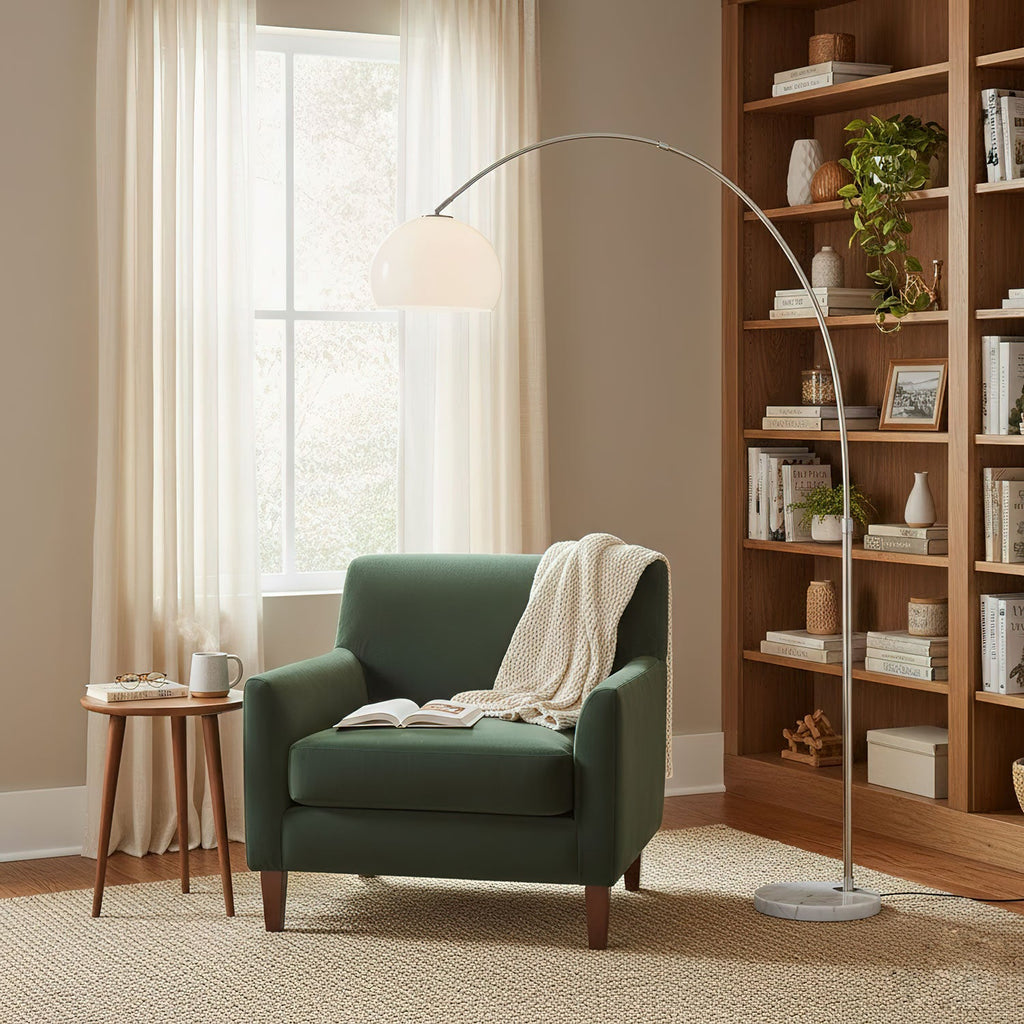 Clérachic arc floor lamp arching over a green armchair with a white throw, beside a wooden bookshelf filled with plants.
