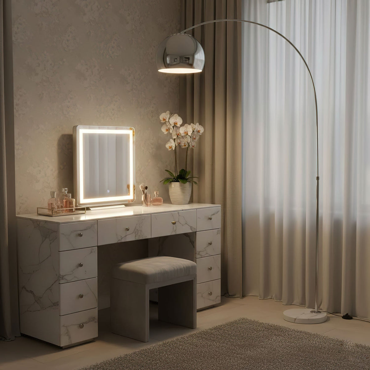 In a bedroom, the Clérachic arc floor lamp's chrome dome shade stands positioned behind a lit marble vanity mirror and an orchid.