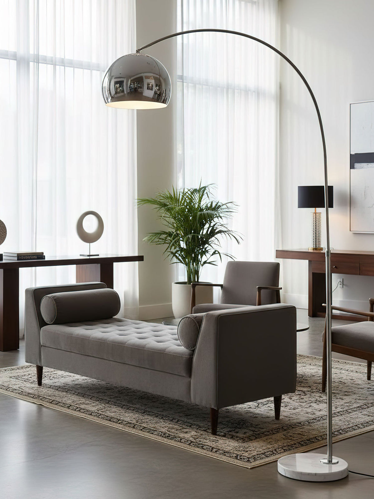 A chrome dome shade tops the Clérachic arc floor lamp as it curves above a grey tufted daybed and a dark side-table lamp.